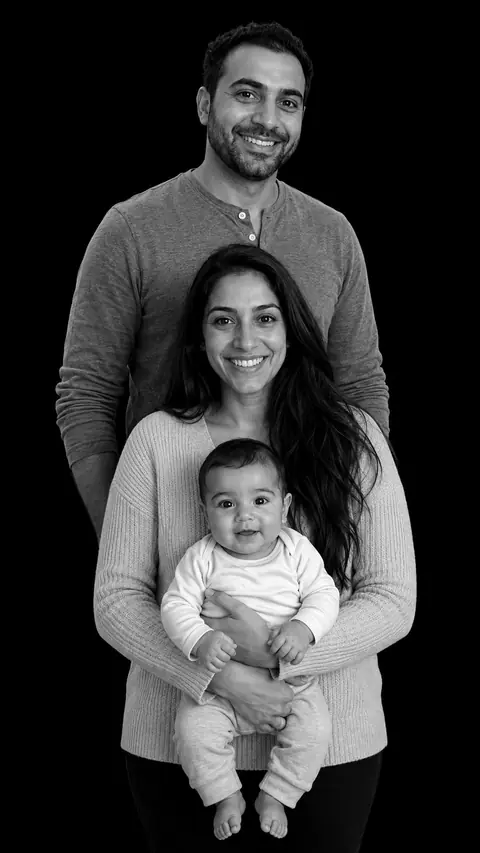 A family of three in a stacked monochrome studio portrait: a man in a henley, a woman with long dark hair, and a baby in a white onesie at the front