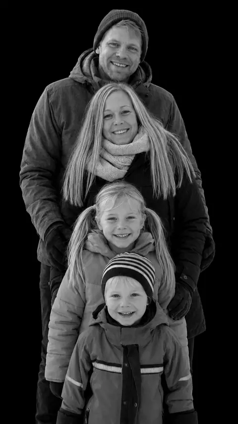 A family of four in winter coats in a stacked monochrome studio portrait, the parents at the back and two fair-haired children in front