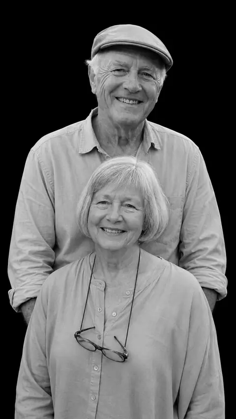An older couple in a stacked monochrome studio portrait: a man in a flat cap behind a woman with a white bob and reading glasses on a cord